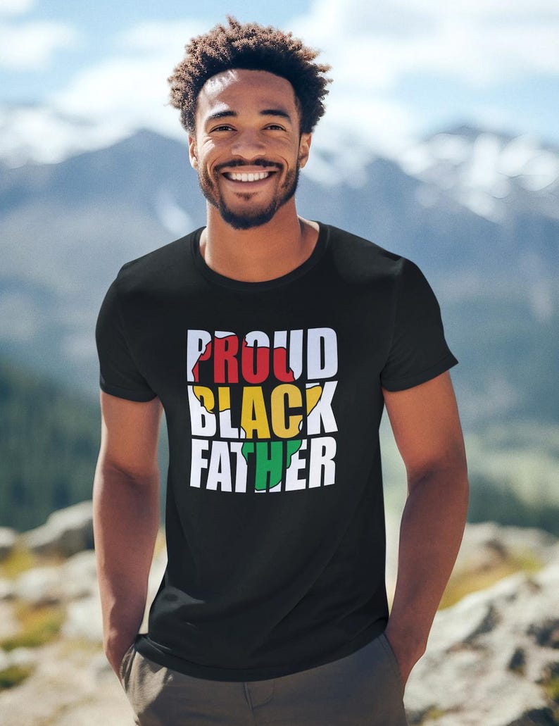 May include: Black t-shirt with the text "PROUD BLACK FATHER" in white, red, yellow, and green. The shirt is worn by a person with a mountain backdrop.