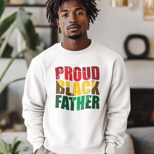 May include: A white sweatshirt with the words "PROUD BLACK FATHER" in red, yellow, brown, and green. The sweatshirt is a crew neck style and is being worn by a person.