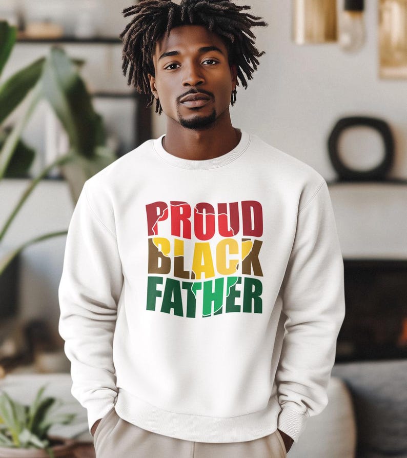 May include: A white sweatshirt with the words "PROUD BLACK FATHER" in red, yellow, brown, and green. The sweatshirt is a crew neck style and is being worn by a person.