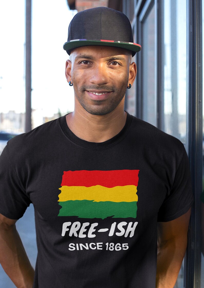 Op de afbeelding: Een zwart t-shirt met een afbeelding van een rood, geel en groen horizontaal gestreepte vlag met een rafelige rand. Onder de vlag staat de witte tekst "FREE-ISH" in een vetgedrukt lettertype, gevolgd door de witte tekst "SINCE 1865" in kleiner lettertype.