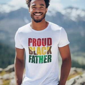 May include: A white t-shirt with the words "PROUD BLACK FATHER" in red, black, yellow, brown, and green. The shirt is worn by a person with a mountain backdrop.