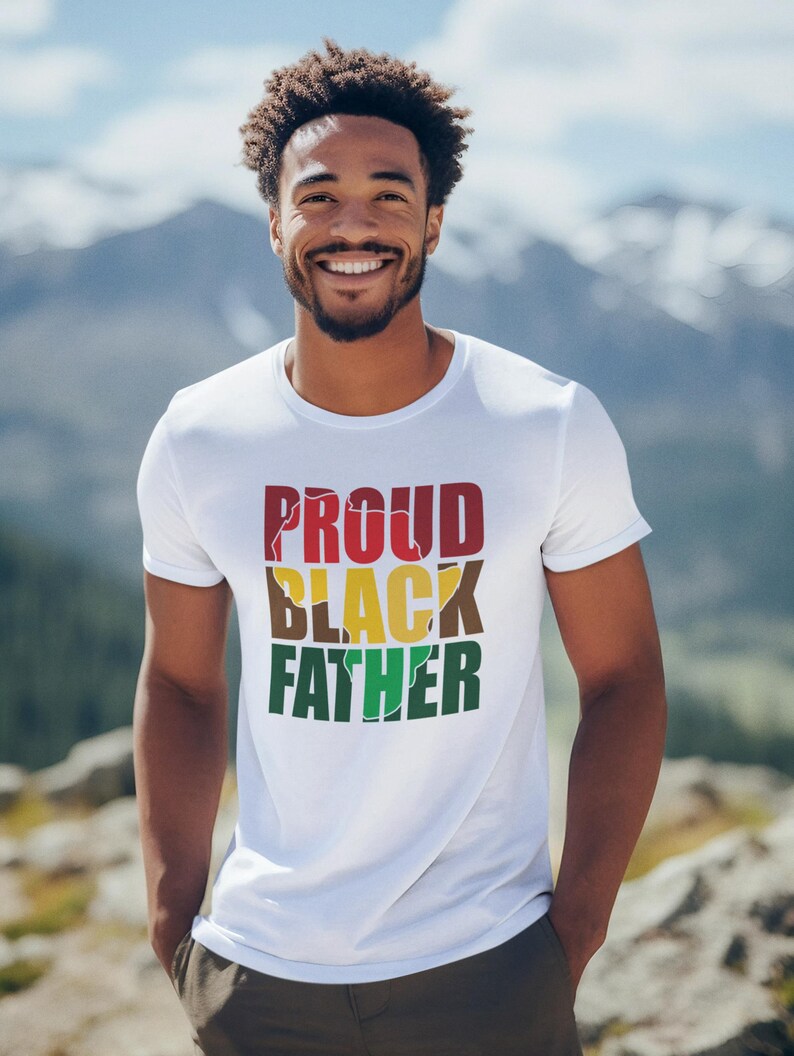 May include: A white t-shirt with the words "PROUD BLACK FATHER" in red, black, yellow, brown, and green. The shirt is worn by a person with a mountain backdrop.