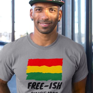 Op de afbeelding: Een grijs T-shirt met korte mouwen en een afbeelding van een rood, geel en groen horizontaal gestreepte vlag met een rafelige rand. Onder de vlag staat de tekst "FREE-ISH" in een vet zwart lettertype, gevolgd door "SINCE 1865" in een kleiner lettertype.