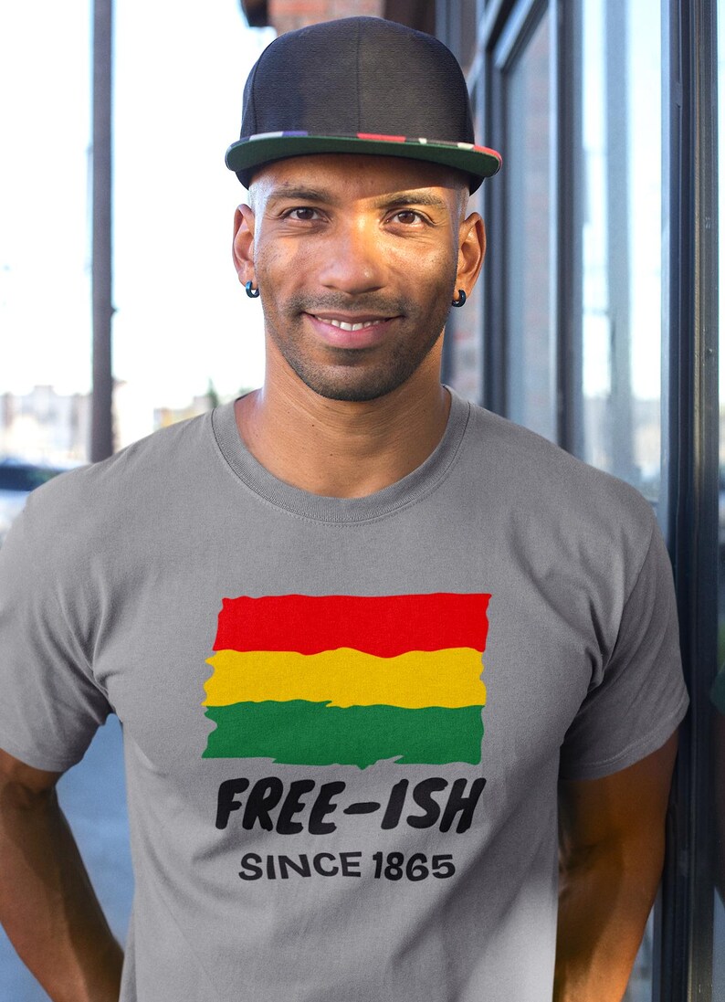 Op de afbeelding: Een grijs T-shirt met korte mouwen en een afbeelding van een rood, geel en groen horizontaal gestreepte vlag met een rafelige rand. Onder de vlag staat de tekst "FREE-ISH" in een vet zwart lettertype, gevolgd door "SINCE 1865" in een kleiner lettertype.