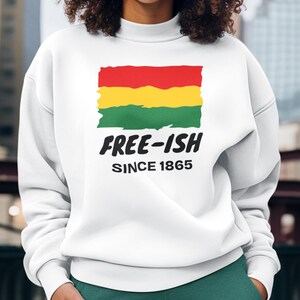 Op de afbeelding: Een wit sweatshirt met lange mouwen en een ronde hals, voorzien van een afbeelding van een vlag met rode, gele en groene penseelstreken. Onder de vlag staat in zwarte, dikke letters "FREE-ISH", gevolgd door de kleinere tekst "SINCE 1865".