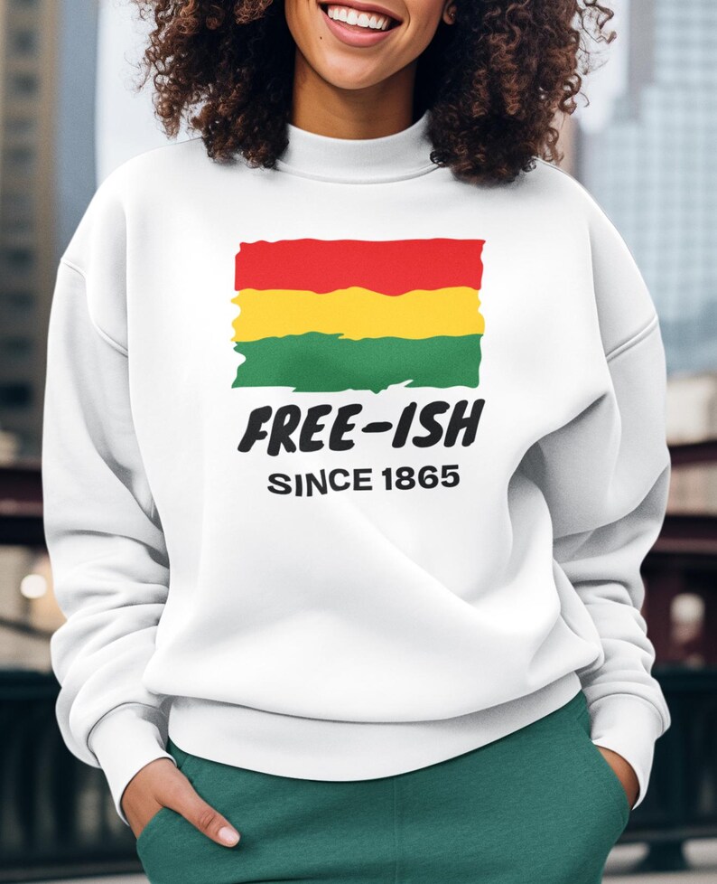 Op de afbeelding: Een wit sweatshirt met lange mouwen en een ronde hals, voorzien van een afbeelding van een vlag met rode, gele en groene penseelstreken. Onder de vlag staat in zwarte, dikke letters "FREE-ISH", gevolgd door de kleinere tekst "SINCE 1865".