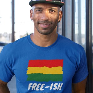 Op de afbeelding: Een koningsblauw T-shirt met korte mouwen en een afbeelding van een rechthoekige vlag in rood, geel en groen met een verweerde textuur. Onder de vlag staat de witte tekst "FREE-ISH" in een vetgedrukt lettertype, gevolgd door "SINCE 1865".