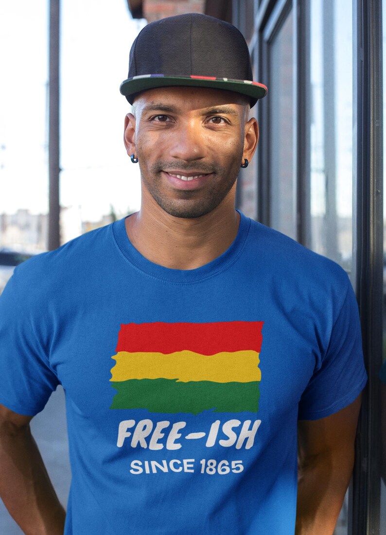Op de afbeelding: Een koningsblauw T-shirt met korte mouwen en een afbeelding van een rechthoekige vlag in rood, geel en groen met een verweerde textuur. Onder de vlag staat de witte tekst "FREE-ISH" in een vetgedrukt lettertype, gevolgd door "SINCE 1865".