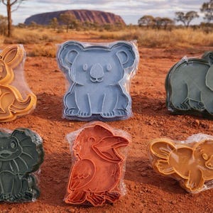 May include: Six plastic cookie cutters shaped like Australian animals: a kangaroo, koala, wombat, frilled-neck lizard, kookaburra, and platypus. The cutters are in different colours and are packaged in clear plastic.