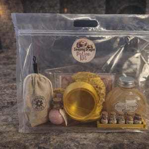 A clear plastic zipper bag containing a gold sleeping dragon potion kit with a cauldron, bottled elixir, and mini jars sitting on a stone surface.