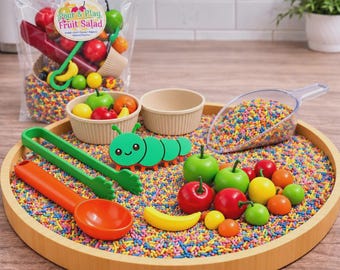 Puede incluir: Un set de juego sensorial con una bandeja redonda de madera llena de chispas de colores, una oruga de plástico verde, una pala, pinzas verdes, una cuchara naranja y frutas de plástico en miniatura como manzanas, cerezas y un plátano.