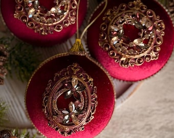 May include: Three round, deep red velvet Christmas ornaments with gold-toned caps and hanging loops. Two ornaments feature circular gold filigree frames with embedded crystals, while the third has an oval frame with floral crystal accents and a red tassel.