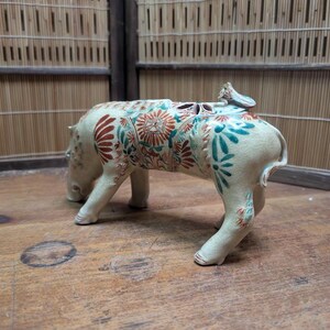 May include: A ceramic hippo figurine with floral patterns in red, teal, and orange. The hippo is cream-colored and has a small bird-like figure on its back. The figurine is on a wooden surface.