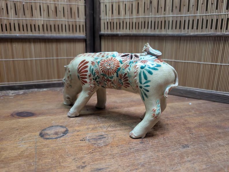 May include: A ceramic hippo figurine with floral patterns in red, teal, and orange. The hippo is cream-colored and has a small bird-like figure on its back. The figurine is on a wooden surface.