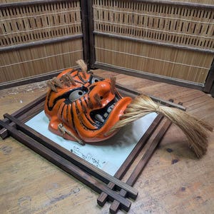 May include: An orange and black ceramic Japanese demon mask with a straw-like element in its mouth. The mask is displayed on a wooden tray with a white surface and calligraphy. The background features a bamboo screen.
