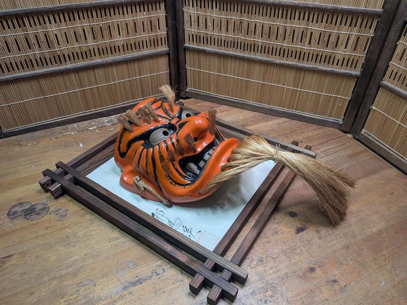 May include: An orange and black ceramic Japanese demon mask with a straw-like element in its mouth. The mask is displayed on a wooden tray with a white surface and calligraphy. The background features a bamboo screen.