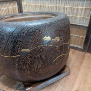 May include: A dark brown wooden bowl with a decorative floral design. The bowl has a rounded shape and sits on a small wooden base. The floral design features gold and blue accents.