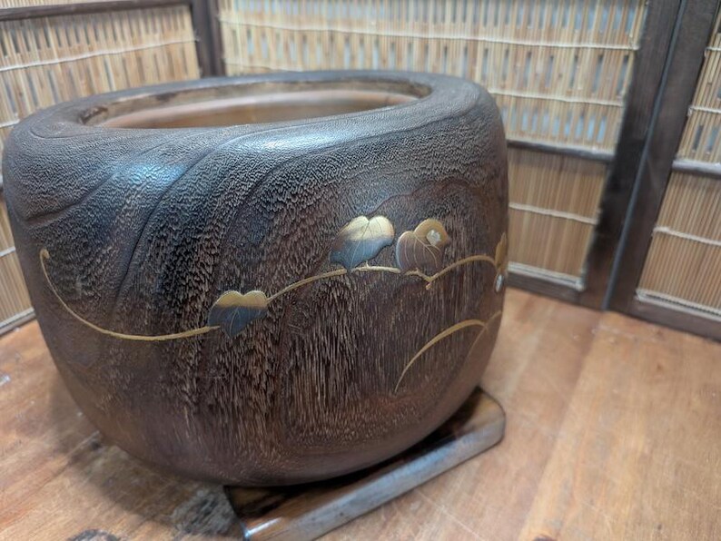 May include: A dark brown wooden bowl with a decorative floral design. The bowl has a rounded shape and sits on a small wooden base. The floral design features gold and blue accents.