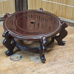 May include: A dark brown, wooden decorative stand with a round top and three curved legs. The stand has a glossy finish and a circular design on the top. It is designed to hold a decorative object.