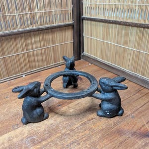 Op de afbeelding: Een decoratieve standaard van gietijzer met drie konijnenfiguren die een cirkelvormige ring ondersteunen. De konijnen zijn donkergrijs van kleur en de standaard staat op een houten oppervlak.