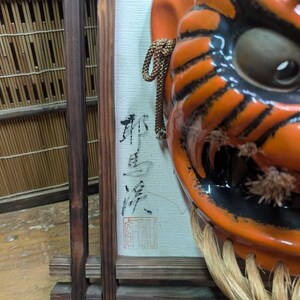 May include: Close-up of a vibrant orange and black Japanese demon mask with a fierce expression. The mask features detailed black stripes, a large eye, and a straw-like beard. A framed artwork with Japanese calligraphy is visible in the background.