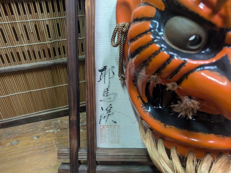 May include: Close-up of a vibrant orange and black Japanese demon mask with a fierce expression. The mask features detailed black stripes, a large eye, and a straw-like beard. A framed artwork with Japanese calligraphy is visible in the background.