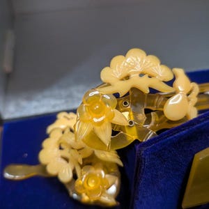 May include: A close-up of a vintage hair comb and hairpin set. The comb and hairpin are a translucent yellow colour, with floral and leaf designs. The set is displayed on a blue velvet surface inside a box.
