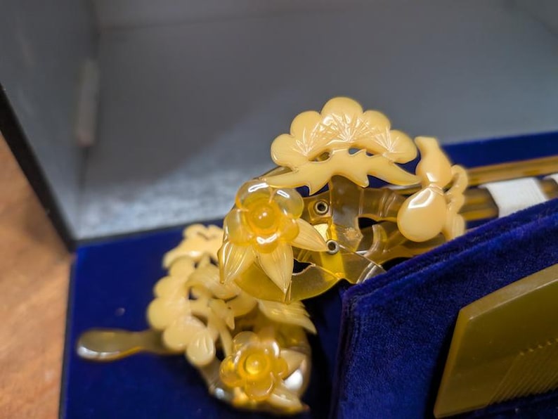 May include: A close-up of a vintage hair comb and hairpin set. The comb and hairpin are a translucent yellow colour, with floral and leaf designs. The set is displayed on a blue velvet surface inside a box.