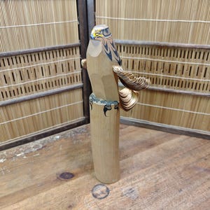 Könnte beinhalten: Eine geschnitzte Holzskulptur eines Falken mit detaillierten Flügeln und einem bemalten Kopf. Der Vogel sitzt auf einem zylindrischen Sockel. Die Skulptur ist aus hellem Holz gefertigt und weist komplizierte Details auf.