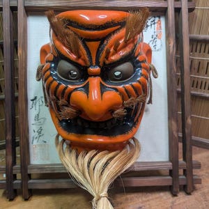 May include: An orange and black Japanese demon mask with a fierce expression. The mask has detailed features, including eyebrows, a nose, and a mouth. It is adorned with a straw-like beard and is set against a framed backdrop.