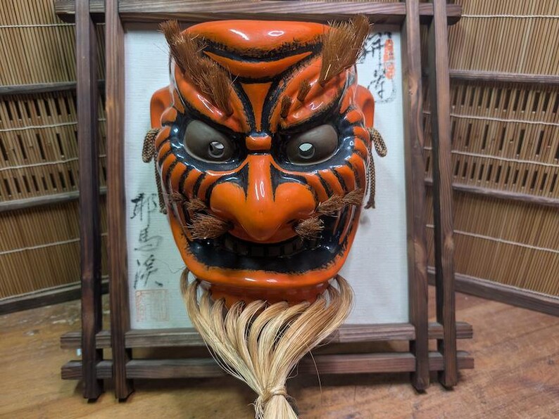 May include: An orange and black Japanese demon mask with a fierce expression. The mask has detailed features, including eyebrows, a nose, and a mouth. It is adorned with a straw-like beard and is set against a framed backdrop.