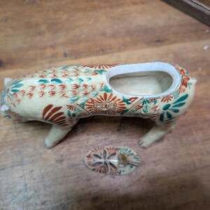 May include: Cream-colored ceramic pig-shaped planter with a rectangular opening and a separate lid. The planter is decorated with red, green, and blue floral patterns. The pig has four legs and a small tail.