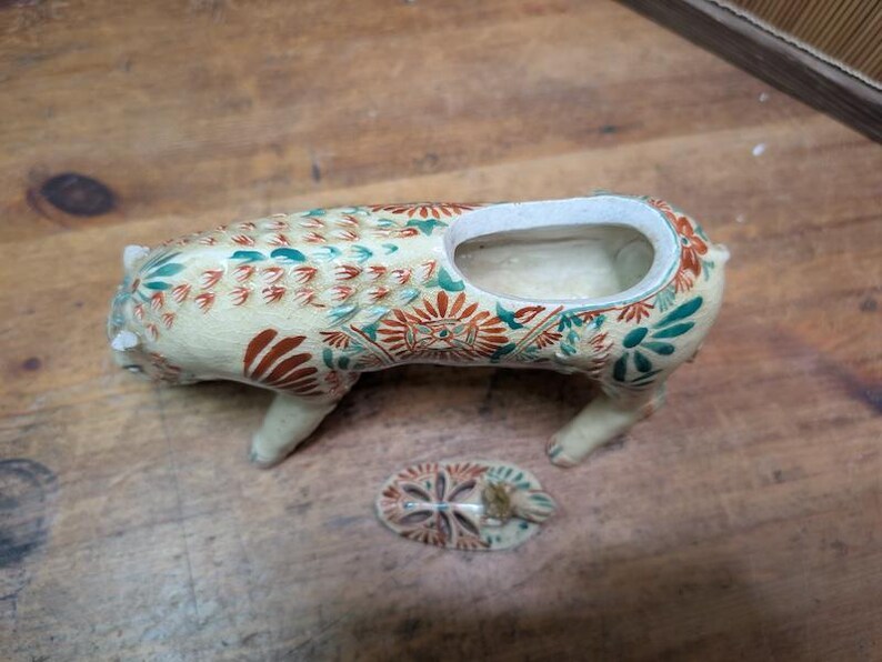 May include: Cream-colored ceramic pig-shaped planter with a rectangular opening and a separate lid. The planter is decorated with red, green, and blue floral patterns. The pig has four legs and a small tail.