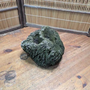 Puede incluir: Una roca texturizada de color verde oscuro con una depresi&oacute;n central, posiblemente un elemento decorativo o natural para la decoraci&oacute;n del hogar. La roca est&aacute; sobre una superficie de madera, con una pantalla de bamb&uacute; al fondo.