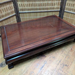 May include: A dark brown wooden table with a rectangular top and a glossy finish. The table has a raised border and rests on four short, curved legs. The wood grain is visible. The table is approximately 120 cm long.