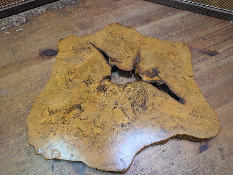 May include: A unique, irregularly shaped wooden slab with a natural edge. The slab is a warm, golden-brown color with darker, intricate grain patterns and a large, dark split. This rustic piece could be used as a decorative element or a small table.