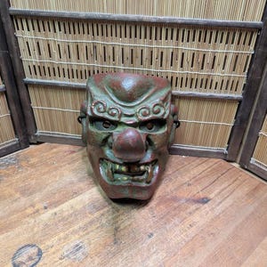 Op de afbeelding: Een verweerde, groen en rood Japanse Noh-masker met een prominente neus en open mond. Het masker heeft gedetailleerde gelaatstrekken en staat tegen een bamboe scherm.