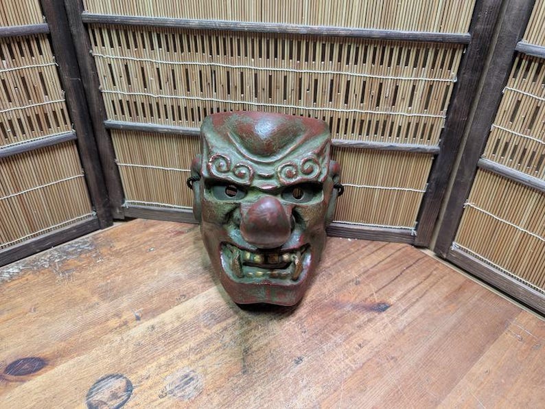 Op de afbeelding: Een verweerde, groen en rood Japanse Noh-masker met een prominente neus en open mond. Het masker heeft gedetailleerde gelaatstrekken en staat tegen een bamboe scherm.