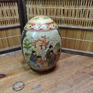 May include: An oval-shaped ceramic decorative egg featuring a cream-colored base with intricate hand-painted scenes of figures in traditional attire near a pavilion, accented by gold detailing, floral motifs, and a patterned border at the top.
