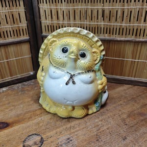 May include: A ceramic Tanuki statue with a yellow hat and body, white belly, and black eyes. The Japanese raccoon dog figurine holds a staff and wears a necklace. The statue is on a wooden surface.