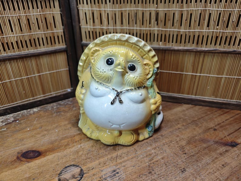 May include: A ceramic Tanuki statue with a yellow hat and body, white belly, and black eyes. The Japanese raccoon dog figurine holds a staff and wears a necklace. The statue is on a wooden surface.