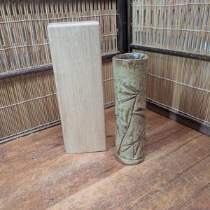 Puede incluir: Un bloque de madera de color claro está junto a un jarrón cilíndrico. El jarrón es de un verde apagado con una superficie texturizada y un diseño de hojas talladas. El fondo presenta una pantalla de bambú.