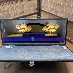 May include: An open black box displaying a decorative item. Inside, a gold-coloured floral design is mounted on a blue velvet base. The box has a handle and a silver clasp.