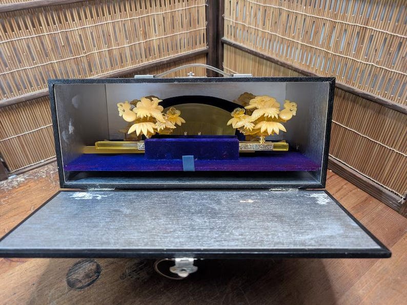 May include: An open black box displaying a decorative item. Inside, a gold-coloured floral design is mounted on a blue velvet base. The box has a handle and a silver clasp.