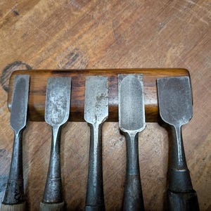 Puede incluir: Un conjunto de cinco cinceles de metal vintage con mangos de madera, dispuestos en una fila sobre un soporte de madera. Los cinceles tienen una apariencia desgastada y envejecida, lo que sugiere que son herramientas antiguas.