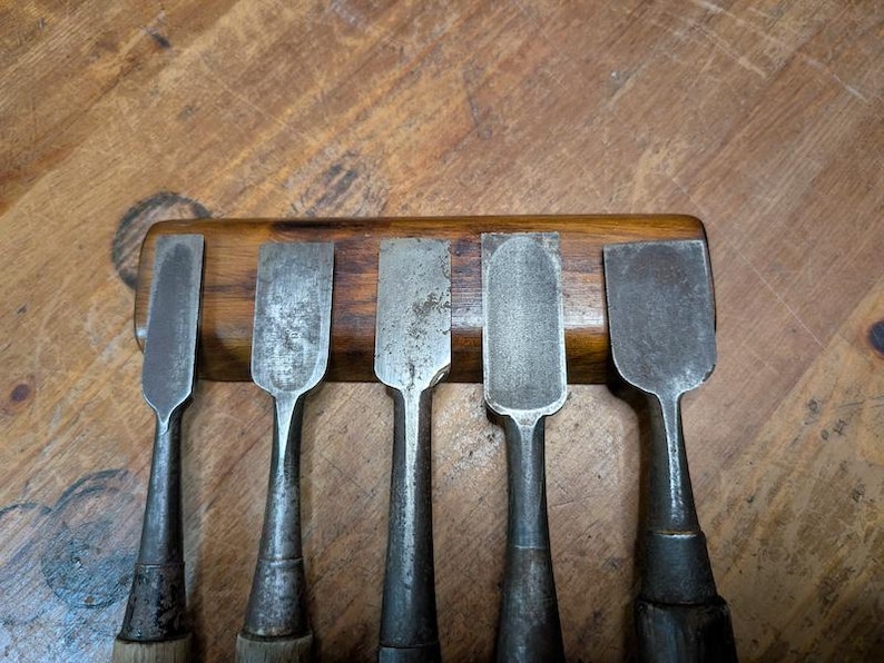 Puede incluir: Un conjunto de cinco cinceles de metal vintage con mangos de madera, dispuestos en una fila sobre un soporte de madera. Los cinceles tienen una apariencia desgastada y envejecida, lo que sugiere que son herramientas antiguas.
