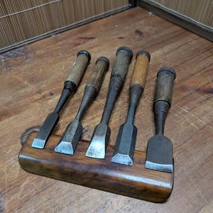 May include: Five vintage wood chisels with wooden handles and metal blades are displayed in a wooden holder. The chisels are arranged in a row, showcasing their different blade shapes and sizes. The wood has a warm, natural tone.