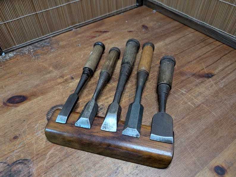 Puede incluir: Cinco cinceles de madera antiguos con mangos de madera y hojas de metal se exhiben en un soporte de madera. Los cinceles est&aacute;n dispuestos en fila, mostrando sus diferentes formas y tama&ntilde;os de hojas. La madera tiene un tono c&aacute;lido y natural.