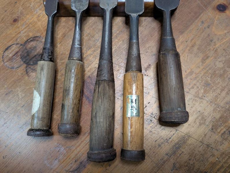 Puede incluir: Colecci&oacute;n de cinco formones de madera antiguos con hojas de metal. Los mangos son de madera envejecida, mostrando signos de desgaste. Los formones est&aacute;n dispuestos sobre una superficie de madera, mostrando su artesan&iacute;a.