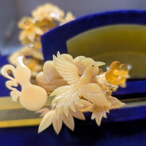 May include: Ivory-coloured hair accessories, including a comb and decorative pieces, are displayed on a blue surface. The comb features a fan shape, while the other pieces showcase intricate bird and floral designs. The accessories are presented in a velvet-lined box.